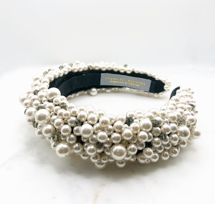 A luxurious black silk velvet headband heavily adorned with high quality white glass pearls and clear and black crystals with a grey label inside and the words FELICITY LEA JEWELLERY HANDMADE IN ENGLAND woven in gold metallic thread. displayed on a marble board