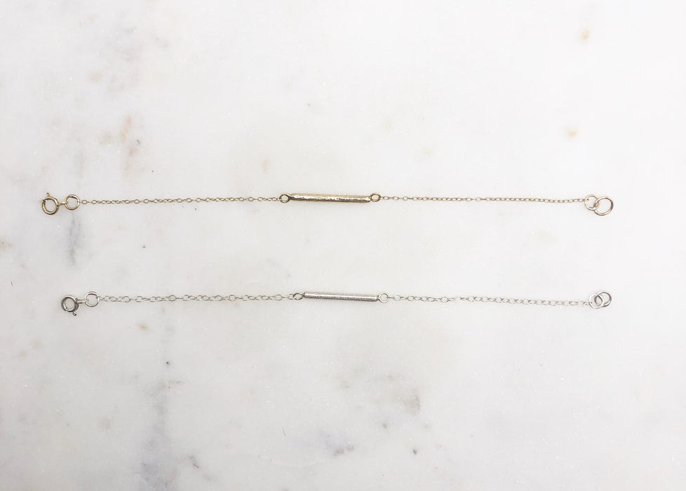 Two bracelets, one gold and one silver displayed on a marble board. Both bracelets feature a textured “twig” bar suspended in the middle of a fine chain
