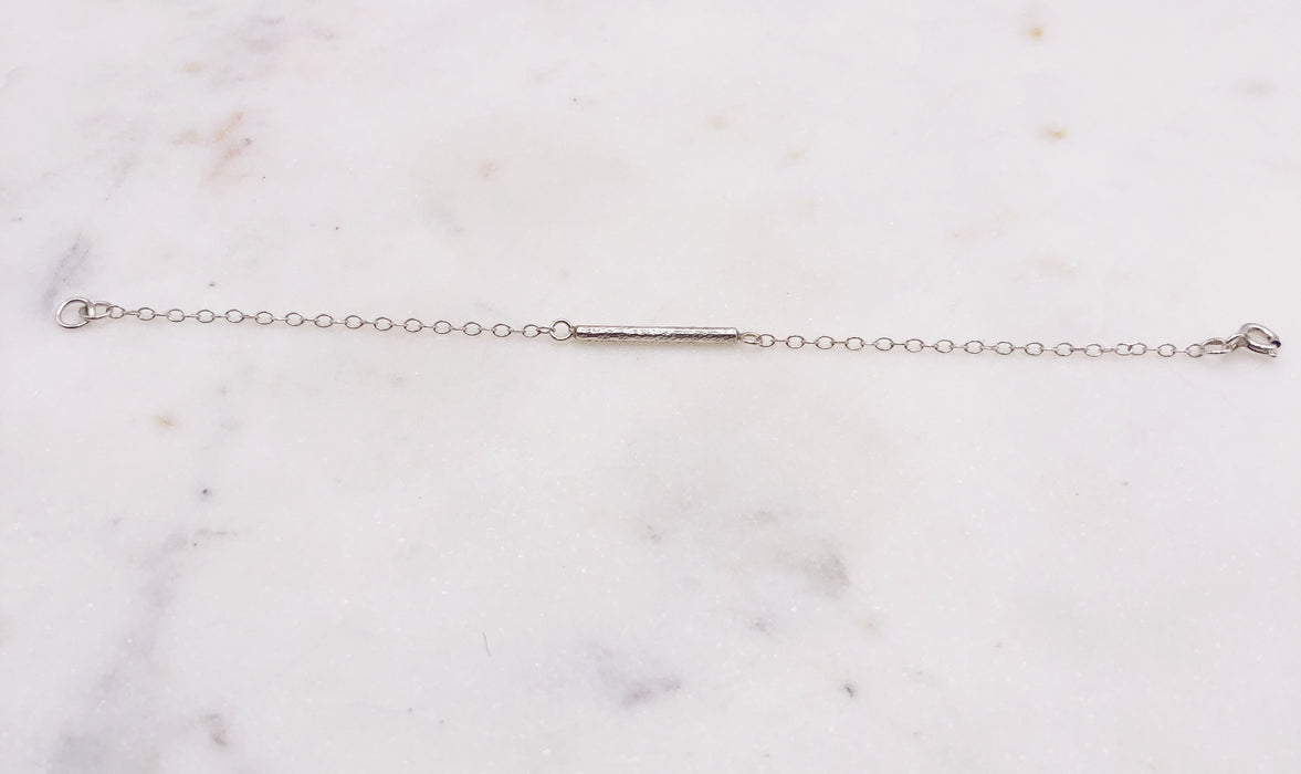 A delicate silver chain bracelet with a decorative solid silver “twig” bar suspended in the middle of the chain and displayed on a marble board.